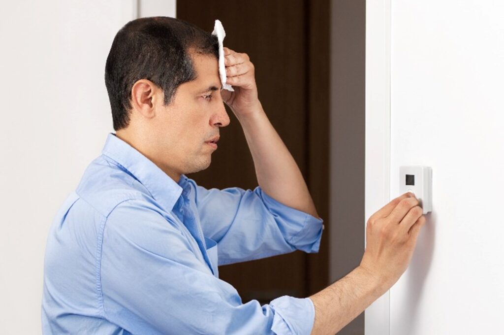 A sweating man looking at a thermostat by Hydro-Flo Plumbing & Heating